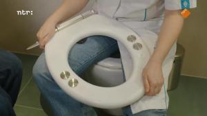 Foto van iemand in een witte jas die een wc-bril op schoot heeft. Het is een witte wc-bril met vier metalen sensoren op de bril. 