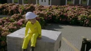 Foto van jongetje in een gele overall en een bouwhelm die zit op een stapel bakstenen