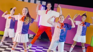 Foto van vier kinderen die samen met danscoach in kleurrijke achtergrond en in kleurrijke kleding een dansje oefenen, ze hebben hun spierballen aangespannen en lachen.