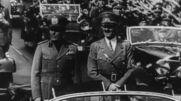 Zwartwitbeeld van Hitler die staand in een auto naast een andere man in legerkleding staat terwijl mensen op straat de Hitlergroet brengen.