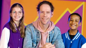 Foto van glimlachende zangcoach Michiel, gekleed in roze hoodie met spijkerjasje, met naast zich twee meisjes van Kinderen voor Kinderen in gelijksoortige paarse en blauwe jacks.