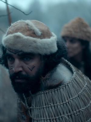 Foto van jagers uit de prehistorie die gespannen kijken naar een prooi. Man met donker haar, baard, bruine muts en tekening op het gezicht loopt voorop.