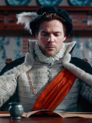 Foto van acteur Thomas Cammaert als Willem van Oranje die achter een tafel zit waarop een document ligt waar hij serieus naar kijkt. In zijn rechterhand houdt hij een witte ganzenveer om het te ondertekenen.