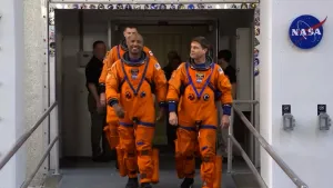 Foto van drie astronauten in oranje pakken die lachend een deur uit komen lopen.