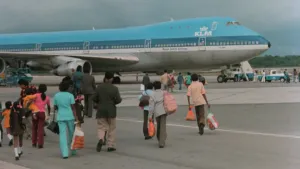 Foto van Surinaamse migranten met koffers en tassen die op het vliegveld naar een blauw KLM-vliegtuig lopen om te migreren naar Nederland.