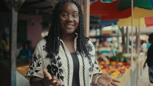 Foto van presentatrice Tirsa in een straat in Suriname. Achter haar staan marktkraampjes met groente, fruit en kleurrijke parasollen.
