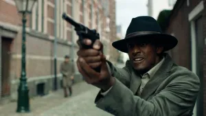 Foto van een donkere man met zwarte hoed die in een straat een ouderwets pistool heft. Hij kijkt gespannen. Zijn slachtoffer is niet zichtbaar. Schuin achter hem loopt nog een man, maar die is onscherp.