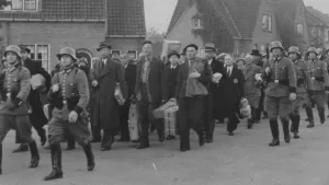 Zwartwitfoto van mannen die in de Tweede Wereldoorlog worden meegenomen door Duitse soldaten. Ze dragen koffers en kisten met spullen.