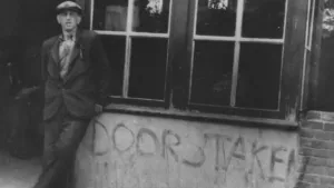 Zwartwitfoto van stakende man met een eenvoudige pet naast een muur waarop geschreven staat 'doorstaken'.