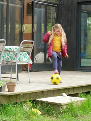 Een meisje met blonde haren staat op het terras bij een huis met haar voet op een gele voetbal.