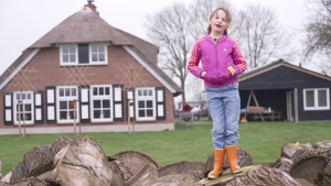 Een meisje staat op een steen, achter haar is haar huis, een boerderij te zien.