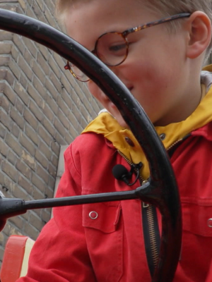 Een foto van een jongetje op een oude rode tractor, het grote stuur zit voor zijn hoofd.