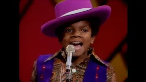 Foto van Michael Jackson als kind tijdens een optreden met de Jackson Five, gekleed in een overhemd met blauw gilet en een paarse hoed schuin op zijn hoofd.