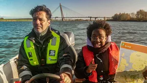 Foto van presentatrice Tirsa Wit op een bootje met naast zich een man aan het stuur. Ze heeft een rood reddingsvest aan en houdt een kaart omhoog van Nederland.
