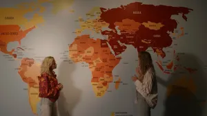 Presentator Janouk en een vrouw staan voor een grote wand met daarop een wereldkaart met gekleurde landen.