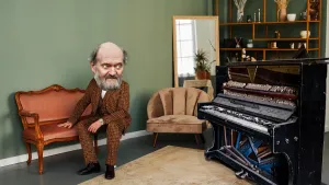 Foto van een kamer met een bank waarop een man met grijze baard zit. Hij kijkt naar een zwarte piano die er een beetje verwaarloosd en kapot uitziet.