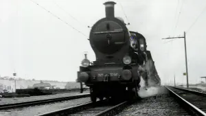 Zwartwitfoto van ouderwetse stoomtrein die over het spoor rijdt. Er komt stoom uit de zijkant van de trein.