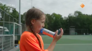 Foto van meisje in oranje sportshirt dat puffertje bij haar mond houdt op het sportveld.