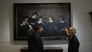 Presentator Nizar en een vrouw staan naast een schilderij van Frans hals met daarop allemaal mannen met hoeden en witte kragen.