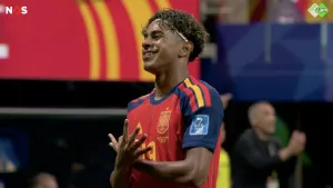 Afbeelding voetballer in een rood‑blauw tenue met gele details op het veld met een handgebaar terwijl op de achtergrond een stadion
