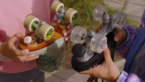 Een foto van twee verschillende rolschaatsen die ondersteboven vastgehouden worden. De ene rolschaats heeft een grotere toestop (rem) dan de andere. De ene rolschaats is lichtgroen, de andere is zwart en heeft doorzichtige wielen met glitters.