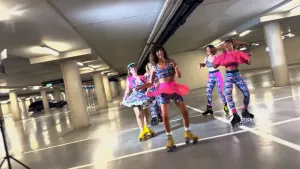 Een foto van vijf danseressen op rolschaatsen. Ze dragen felgekleurde kleding in blauw en roze en staan op de gladde vloer van een parkeergarage.