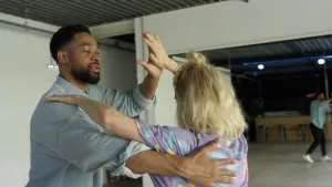 Een foto van Janouk die salsadansles krijgt van een dansleraar. Ze dansen samen tegenover elkaar in stijldanshouding, hij heeft zijn arm op haar rug gelegd en leidt met zijn andere hand. Janouk houdt haar hand tegen die hand. Ze dansen in een dansleszaal.