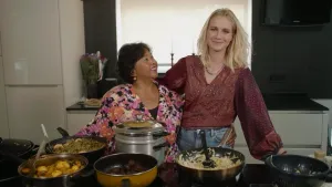 Presentatrice Janouk staat naast een vrouw met Indonesische roots in een keuken. Voor hen staan allemaal pannen met eten erin.