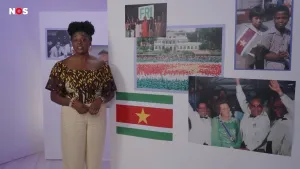 Een foto van een presentatrice in een studio. Naast haar staat een fotowand met de vlag van Suriname erop, en een foto van Koningin Beatrix met Surinaamse mannen, en kinderen met een Surinaams vlaggetje.