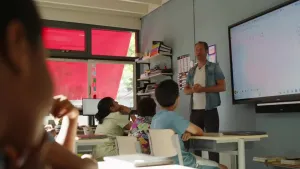 Foto van een klaslokaal waarin een docent voor een digitaal bord staat en lesgeeft aan zittende leerlingen terwijl zonlicht door ramen met rode zonwering naar binnen valt.