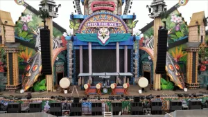 Foto van vooraanzicht festivalpodium in junglethema met centraal de draaitafel van de dj, links en rechts grote zwarte speakers en grote trommels op het podium.