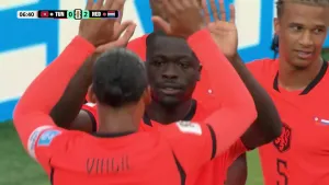 Foto van voetballers in oranje shirts die elkaar high-fives geven tijdens een wedstrijd, met linksboven een scorebord waarop Tunesië 0 en Nederland 2 staat.