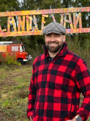 Een foto van boswachter Tim in een zwart-rood geruit overhemd voor een bord met de tekst ' Het landje van Tim'.