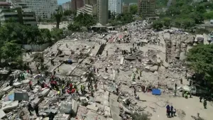 foto van een verwoest stadsgebied met ingestorte gebouwen en veel puin, waar reddingswerkers en omstanders tussen de brokstukken zoeken naar overlevenden.