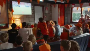 Foto van een groep kinderen en jongeren in oranje kleding en accessoires die binnen naar een geprojecteerde voetbalwedstrijd kijken.