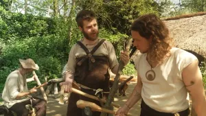 Een foto van een man in kleding uit de tijd van jagers en boeren: een simpel t-shirt en een soort beschermende rok die hoog tot boven de borst komt. Hij draagt bronzen sieraden en laat een bijl zien aan presentator Pascal.