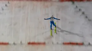 Een foto van een schansspringer, een man op ski's in een strak blauw pak met een helm op. Hij komt op hoge snelheid neer op een besneeuwde helling. Horizontaal loopt een rode streepjeslijn over de helling.