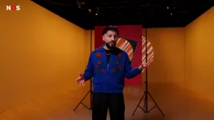 Een foto van presentator Devran in de studio. Hij houdt zijn armen uit elkaar, als een soort weegschaal. Achter hem hangt een poster van een groot stemvakje dat rood ingekleurd is. De studio is gekleurd met geel licht.