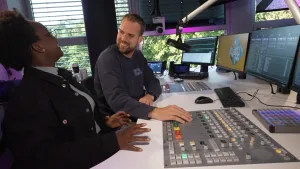 Een foto van radio-dj Wijnand Speelman en Klokhuis-presentatrice Tirsa in een radiostudio. Wijnand heeft zijn hand op een mengpaneel met knoppen, voor zijn gezicht hangt een microfoon. Er staan vier computerschermen.