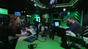 Een foto van een radiostudio. Rechts zit de dj, Wijnand. In het midden regisseur Joerie. Links zitten redacteur Lideke en samensteller Marc. Ze zitten aan een grote ovale tafel met veel beeldschermen en microfoons.