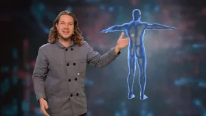 Een foto van presentator Pascal in een studio, hij kijkt in de camera. Naast hem zweeft een blauwgekleurd hologram van een menselijk lichaam, een soort projectie van een afbeelding. Door het hologram heen is de hand van Pascal te zien.