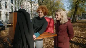 Een foto van twee vrouwen die buiten bij een grote, ouderwetse fotocamera staan.  De camera is van hout en is net iets groter dan een krat. De ene vrouw bedient de camera, presentatrice Janouk kijkt toe.