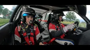 Een foto van twee mensen in een auto. Ze dragen allebei beschermende racepakken en helmen. Ze kijken geconcentreerd naar de weg.