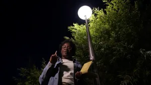 Een foto van presentatrice Tirsa onder een straatlantaarn die aanstaat, het is nacht. Ze wijst naar de lantaarn en heeft een boek in haar hand.