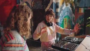 Een foto van een vrouw met een koptelefoon om haar nek, dj Isis. Ze praat met presentator Pascal. Ze staat bij een dj-set, een plat vierkant apparaat met veel knopjes en schuifjes en twee grote draaischijven.