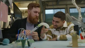 Een foto van presentator Nizar en een man met een volle rode baard. De man houdt een spuit gevuld met lucht vast, hij spuit lucht in een flexibele arm van een robot die eruit ziet als een gekromde vinger. Nizar kijkt geïnteresseerd en wijst ernaar.