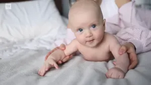 Een foto van een blote baby die op een bed aan het kruipen is. De baby wordt bij de armpjes vastgehouden door iemand erachter.