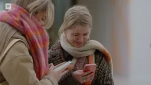 Een foto van twee jonge blonde meiden die allebei op hun telefoon kijken. Ze hebben jassen aan en kleurige sjaals om.