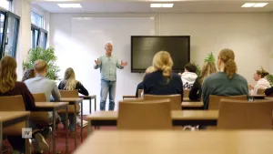 Een foto van een klaslokaal van achterin gezien. Voor de klas staat een man in een mintgroene blouse, hij legt iets uit aan de leerlingen, ze zijn rond de 16 jaar oud.