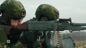 Een foto van twee jonge dienstplichtigen die getraind worden in het leger. Ze liggen naast elkaar op hun buik, eentje schiet met een machinegeweer. Ze dragen groene camouflage-kleding en groene helmen.